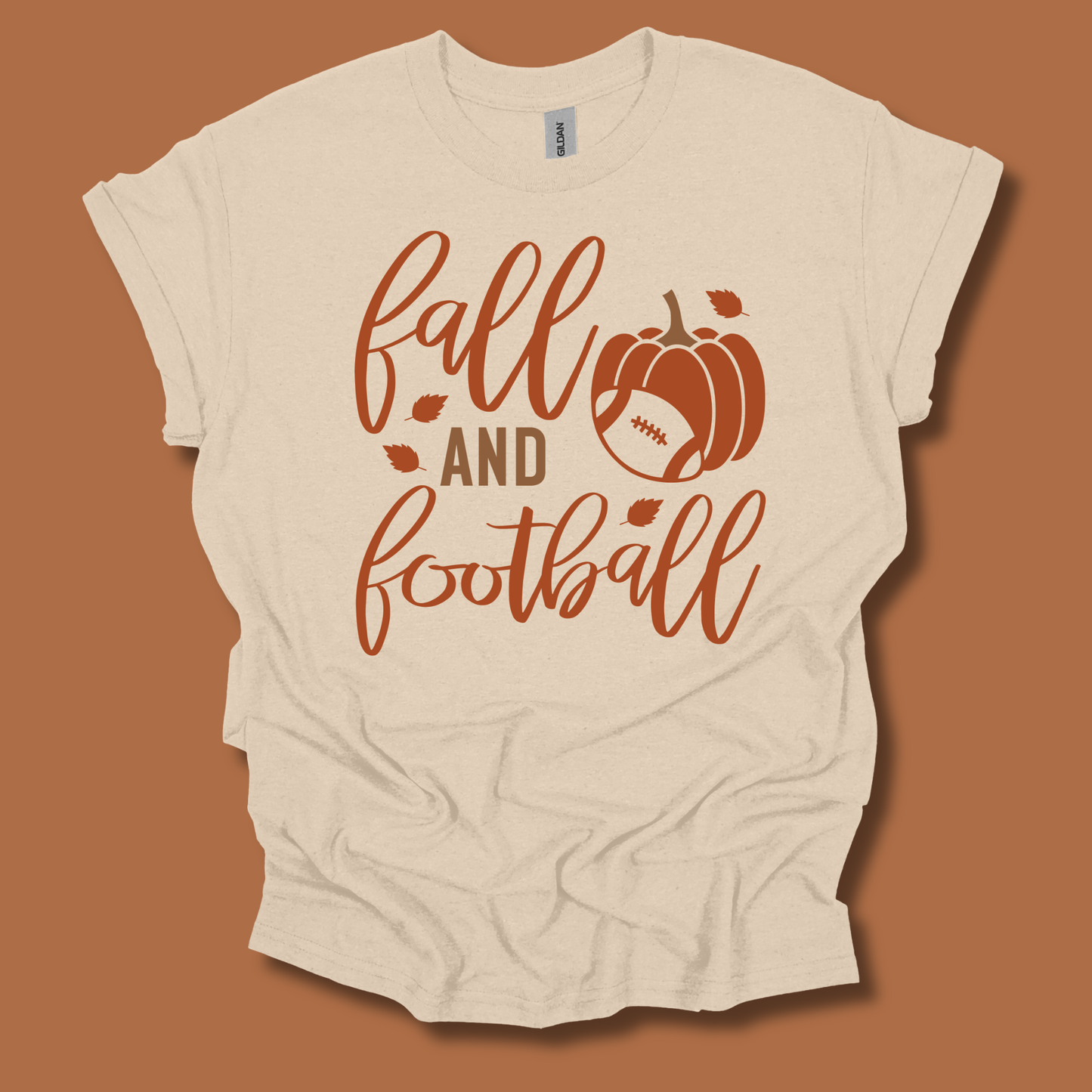 A cream-colored t-shirt with a 'fall and football' graphic in rust orange, featuring a pumpkin and football design, laid flat against a solid rust-brown background.