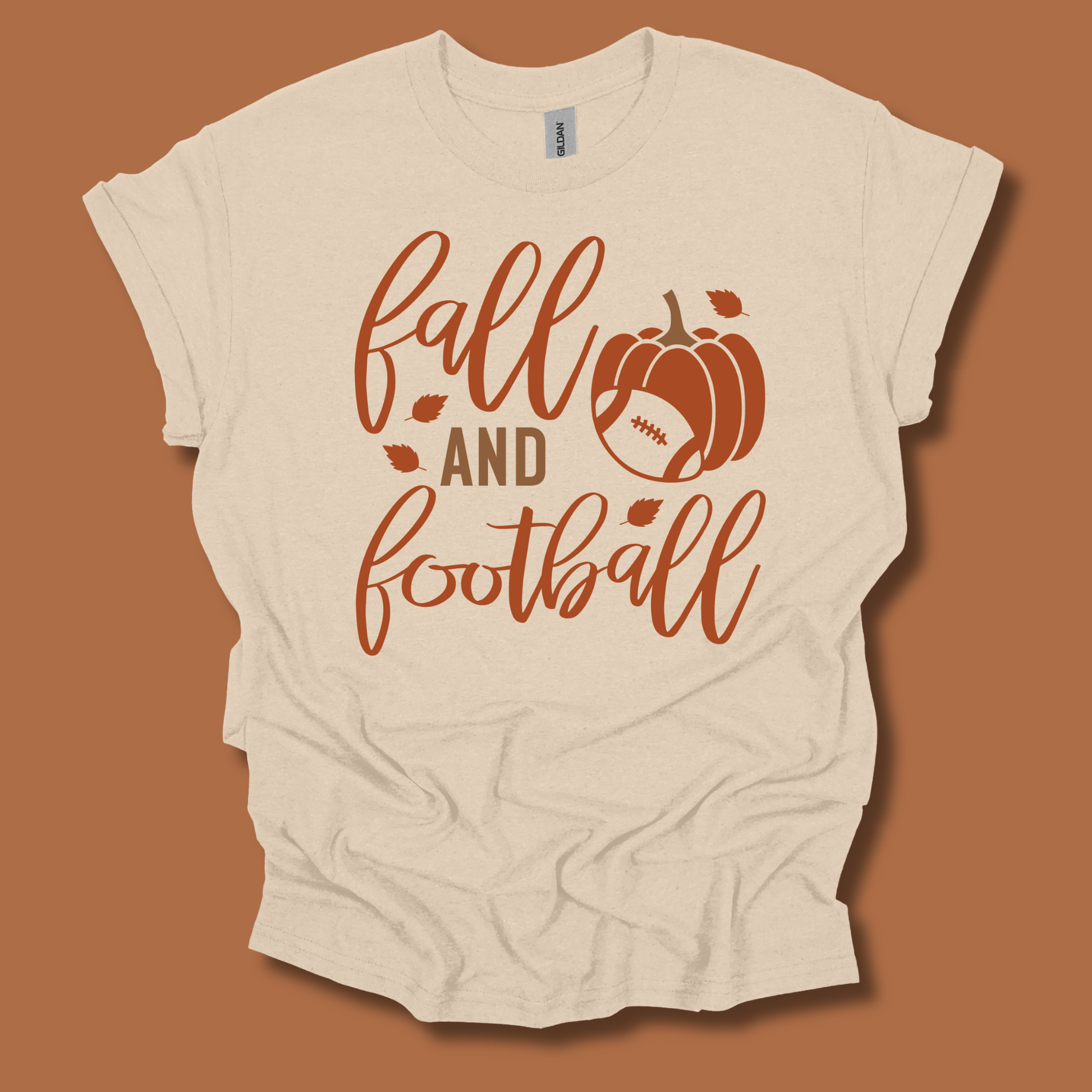 A cream-colored t-shirt with a 'fall and football' graphic in rust orange, featuring a pumpkin and football design, laid flat against a solid rust-brown background.