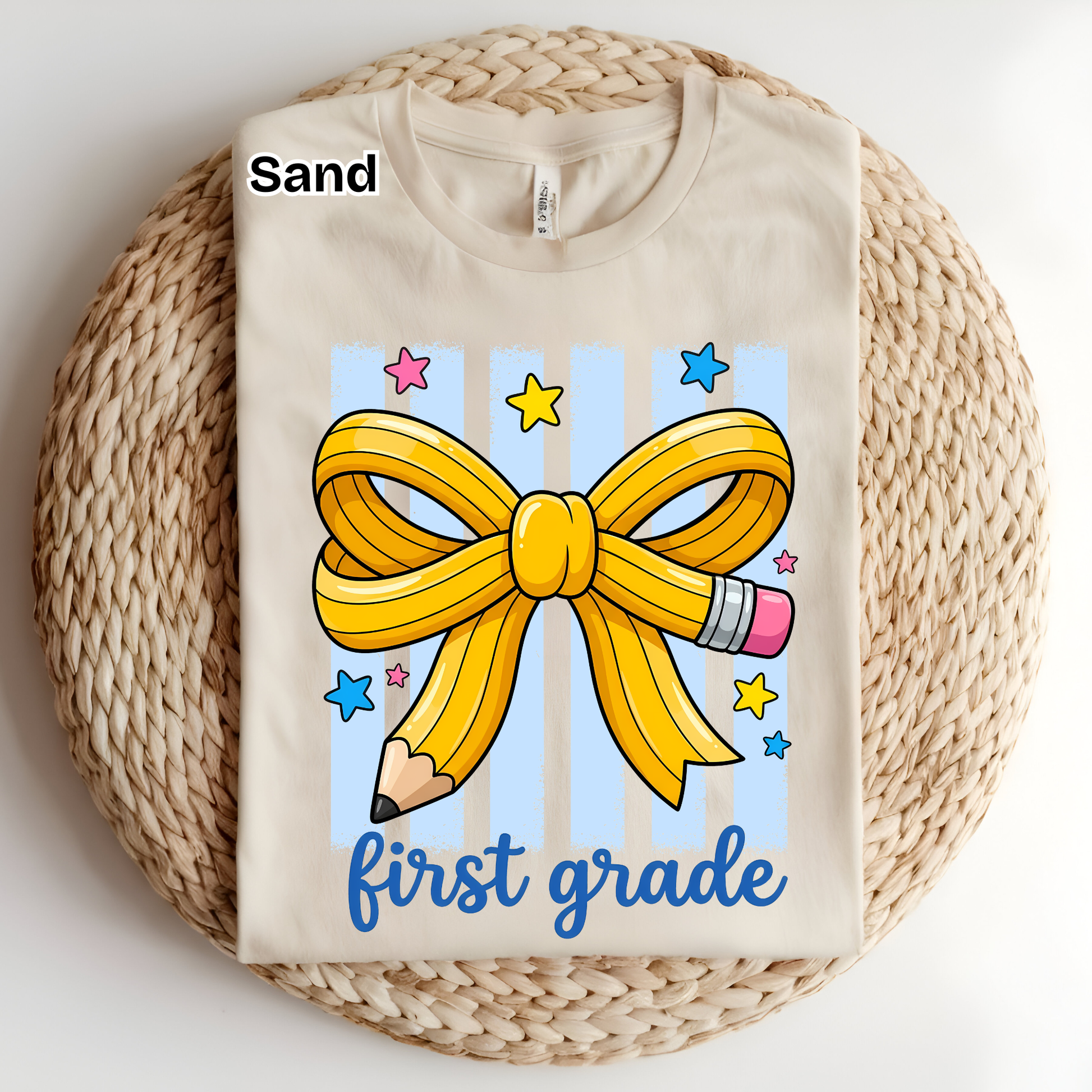 A folded sand-colored t-shirt displayed on a round woven straw mat. The shirt features a graphic design with a yellow ribbon bow, a pencil, blue stars, and the text 'first grade' in blue script.