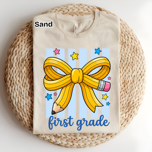 A folded sand-colored t-shirt displayed on a round woven straw mat. The shirt features a graphic design with a yellow ribbon bow, a pencil, blue stars, and the text 'first grade' in blue script.