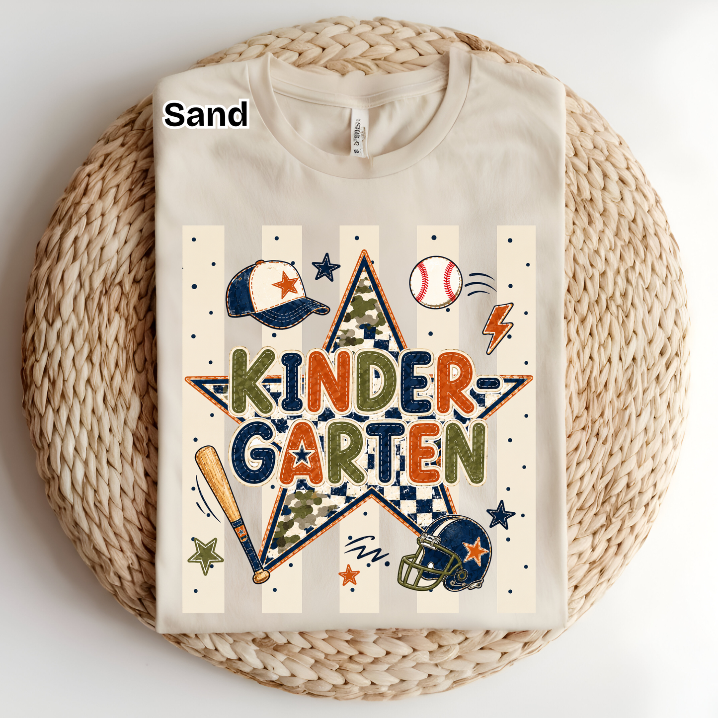A folded cream-colored t-shirt with a 'Kinder Garten' baseball graphic, displayed on a round woven straw mat. The graphic features a star with camouflage and checkered patterns, sports items like a baseball, bat, and helmet, and the word 'SAND' is printed on the collar label.