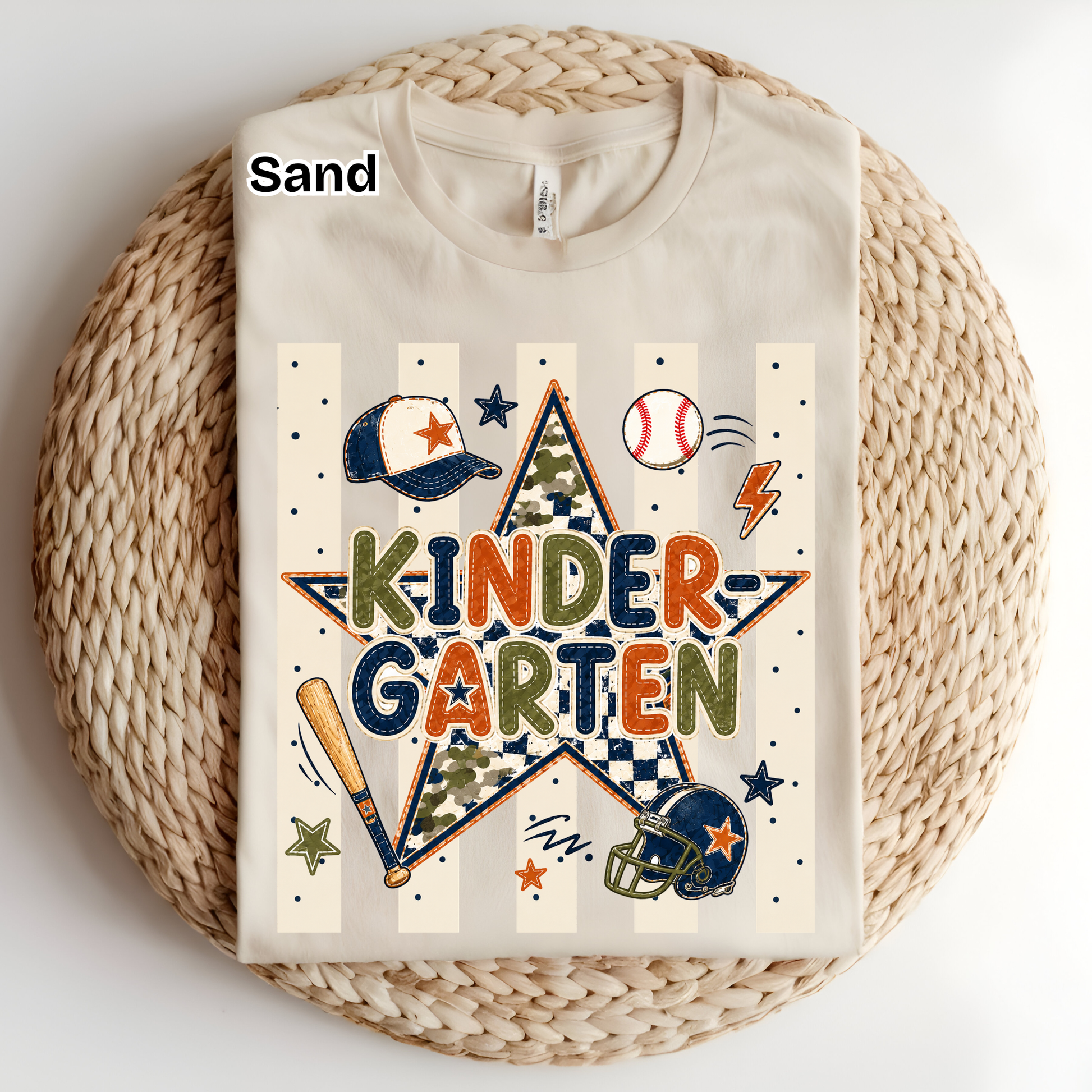 A folded cream-colored t-shirt with a 'Kinder Garten' baseball graphic, displayed on a round woven straw mat. The graphic features a star with camouflage and checkered patterns, sports items like a baseball, bat, and helmet, and the word 'SAND' is printed on the collar label.