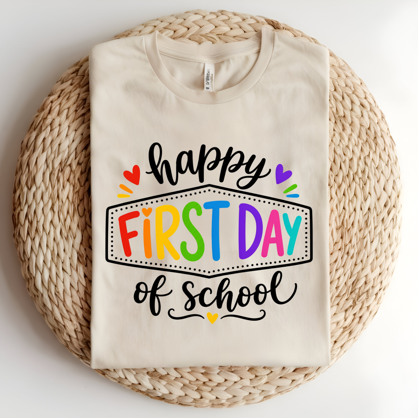 A folded cream-colored t-shirt with a colorful 'happy FIRST DAY of school' graphic, displayed on a round woven straw mat against a white background.