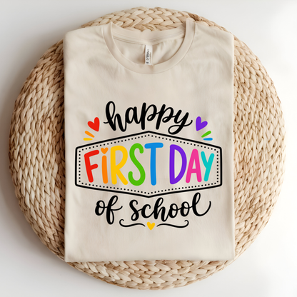 A folded cream-colored t-shirt with a colorful 'happy FIRST DAY of school' graphic, displayed on a round woven straw mat against a white background.