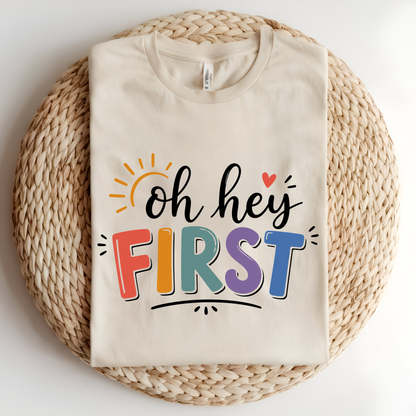 A folded cream-colored t-shirt with a colorful 'Kinder Garten' graphic print, displayed on a round woven straw mat against a white background.