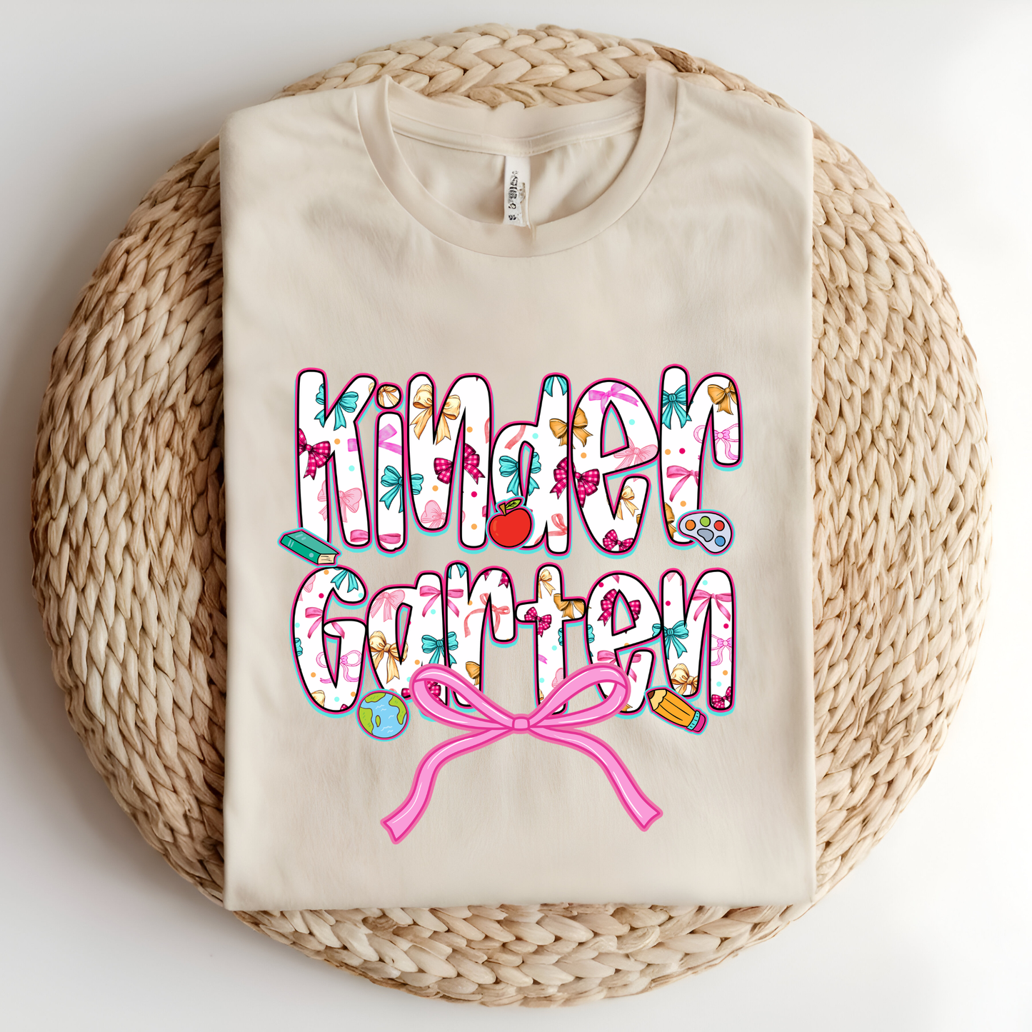 A folded cream-colored t-shirt with a colorful 'Kinder Garten' graphic print, displayed flat on a round woven straw mat against a white background.