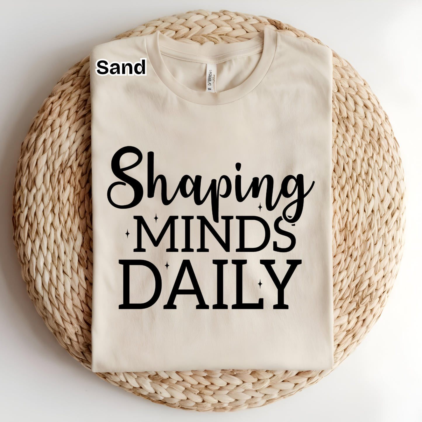 A folded cream-colored t-shirt with the text 'Shaping MINDS DAILY' in black, displayed on a round woven straw mat. The word 'Sand' is printed in black near the collar, indicating the color name.