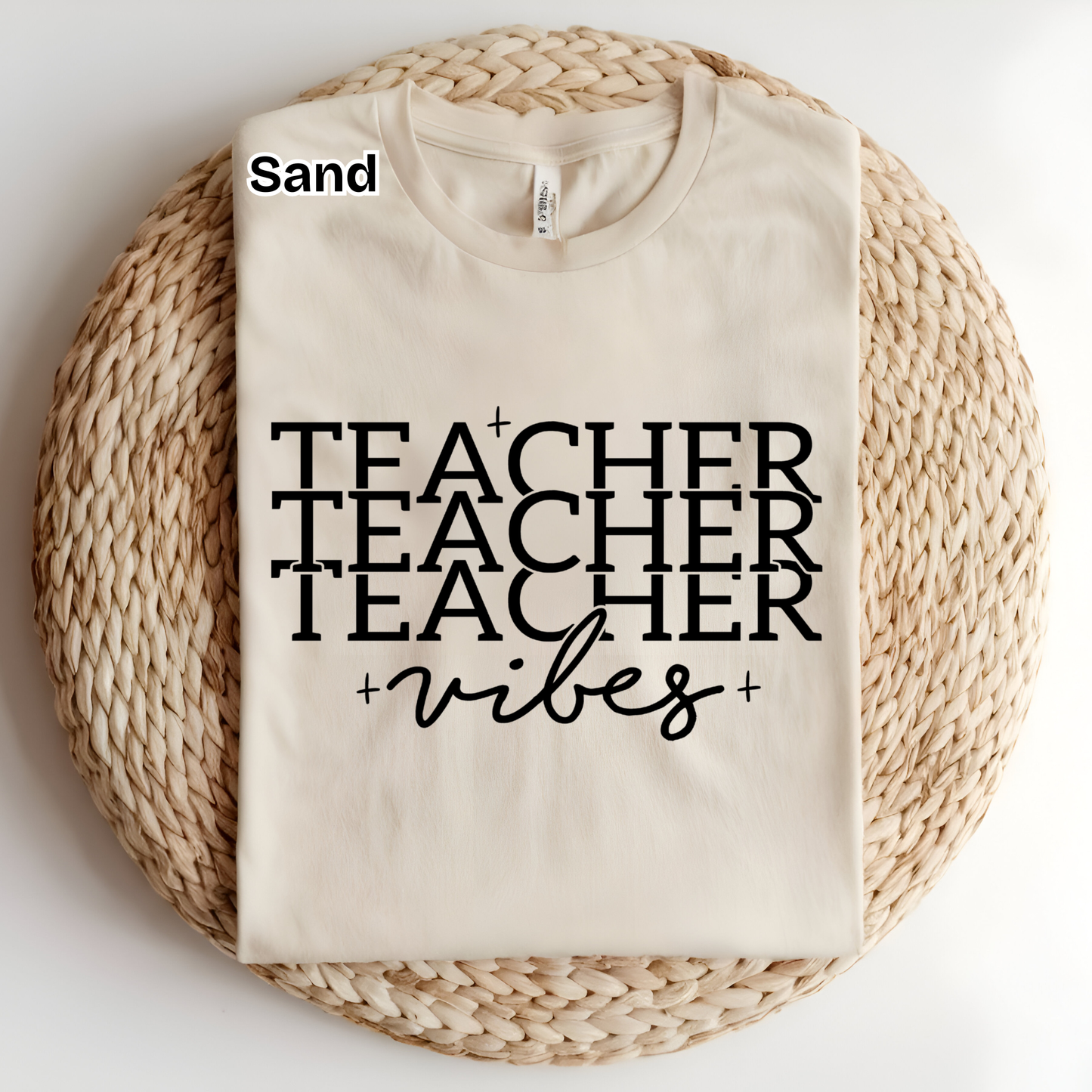 A folded cream-colored t-shirt with black text reading 'TEACHER TEACHER TEACHER +vibes+' is displayed on a round woven straw mat. The word 'Sand' is printed in black near the collar, indicating the fabric color.