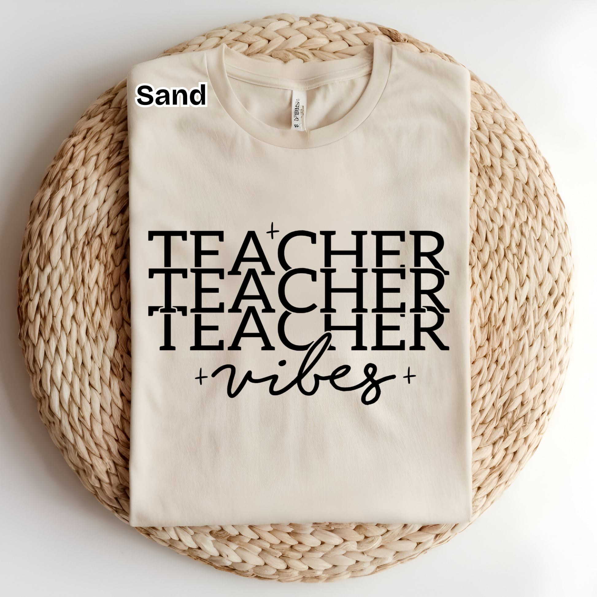 A folded cream-colored t-shirt with black text reading 'TEACHER TEACHER TEACHER +vibes+' is displayed on a round woven straw mat. The word 'Sand' is printed in black near the collar, indicating the fabric color.