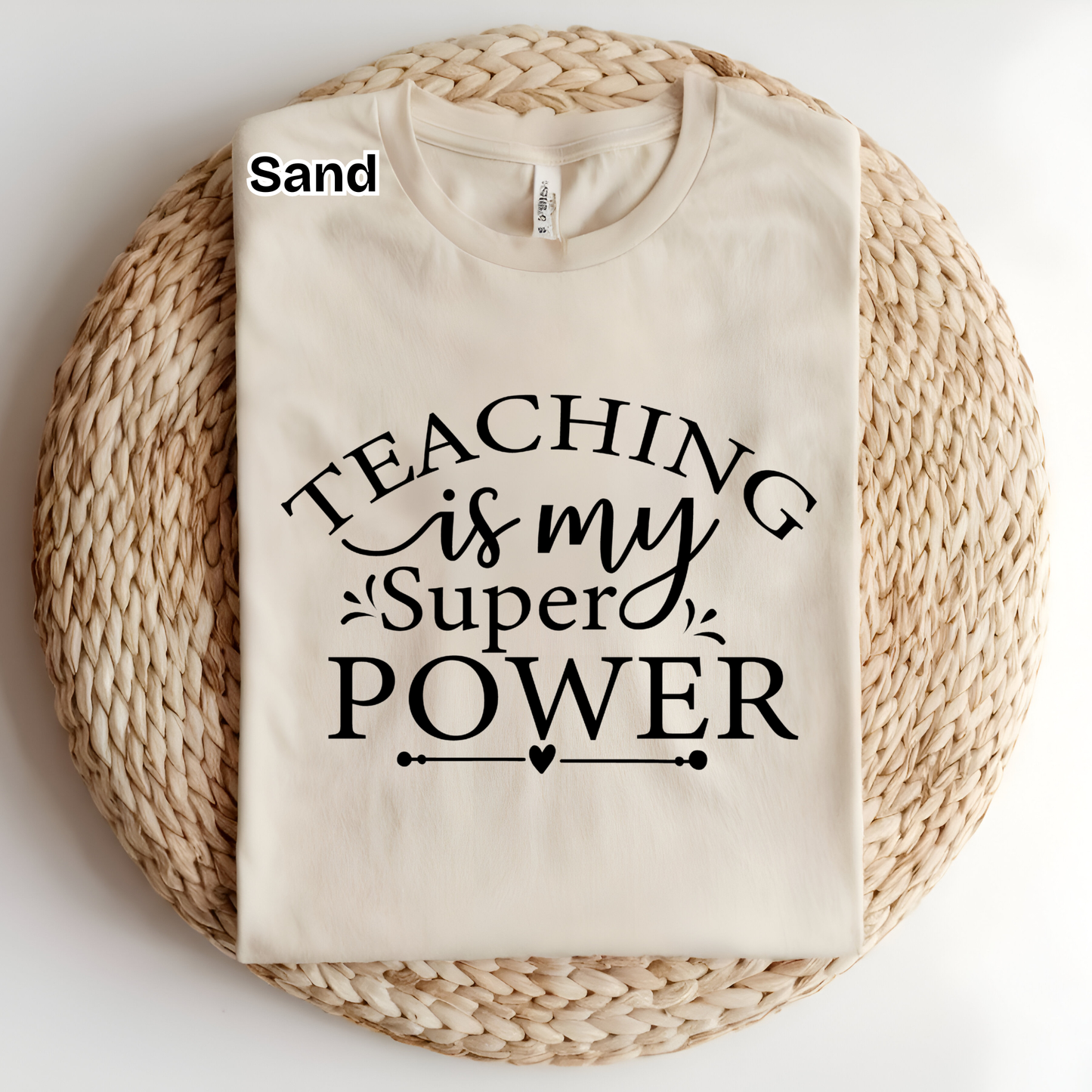 A folded cream-colored t-shirt with black text reading 'TEACHING is my Super POWER' is displayed on a round woven straw mat. The word 'Sand' is printed in black in the top-left corner of the image.