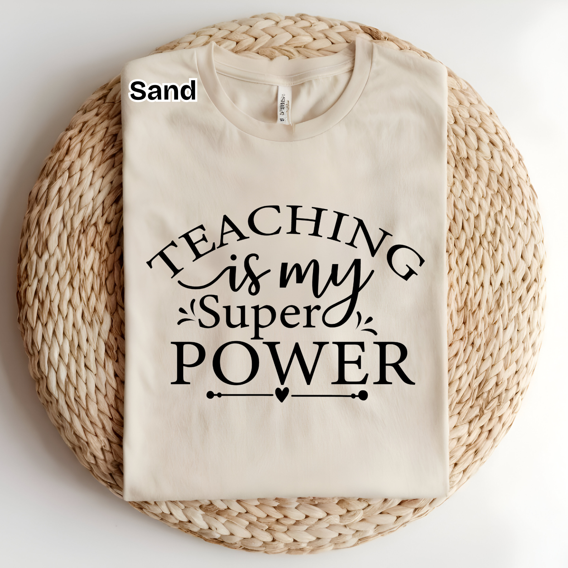 A folded cream-colored t-shirt with black text reading 'TEACHING is my Super POWER' is displayed on a round woven straw mat. The word 'Sand' is printed in black in the top-left corner of the image.