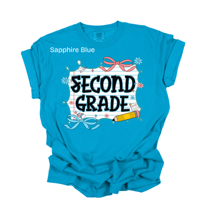 A bright blue short-sleeve t-shirt with a 'SECOND GRADE' graphic on the front, featuring a white scroll, black text, a pink bow, and a yellow pencil. The text 'Sapphire Blue' is printed on the upper left chest.