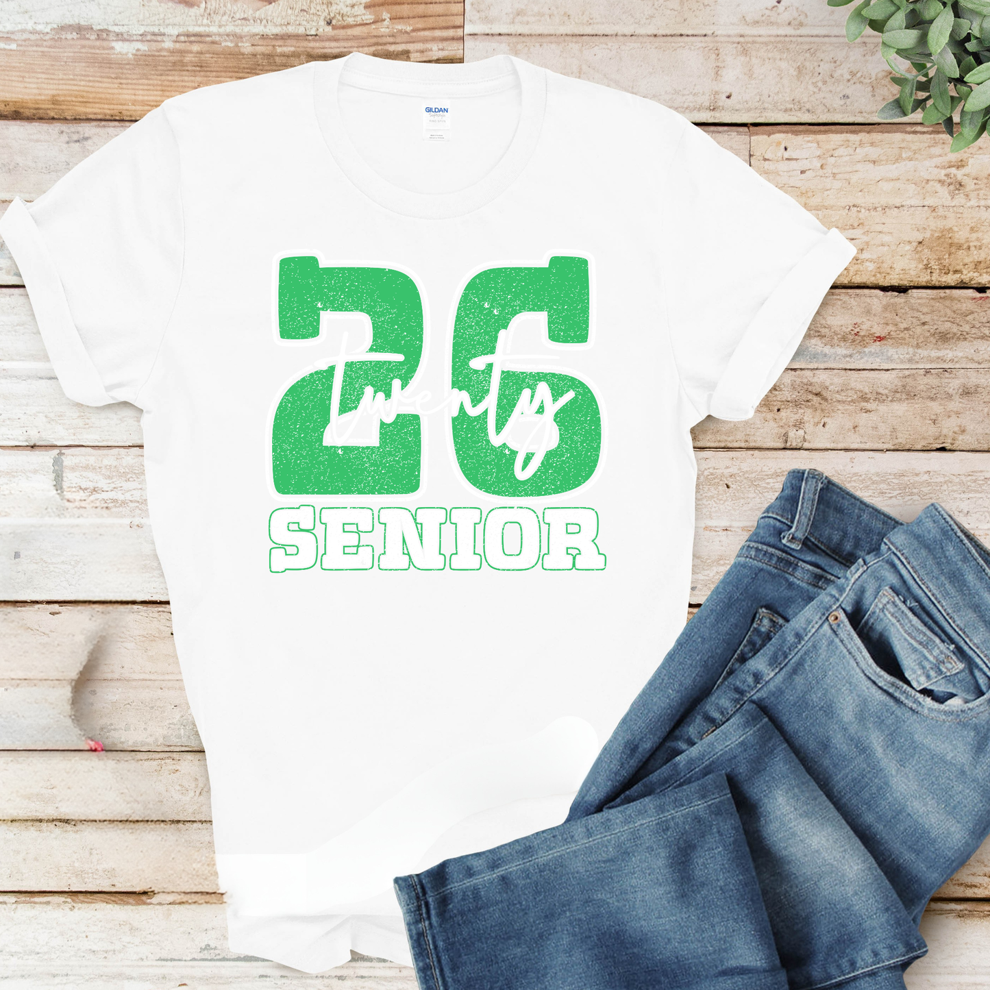 White t-shirt with green '26 Senior' design on a wooden surface with jeans.