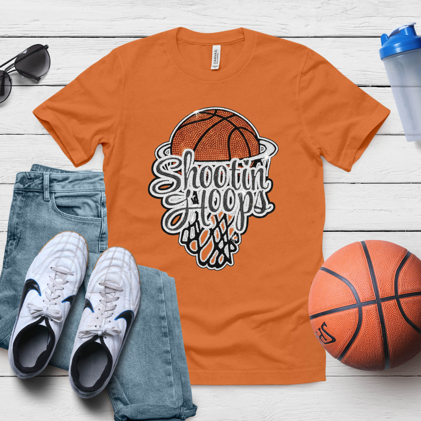 Orange t-shirt with 'Shooting Hoops' graphic, white sneakers, blue jeans, and a basketball on a wooden surface.