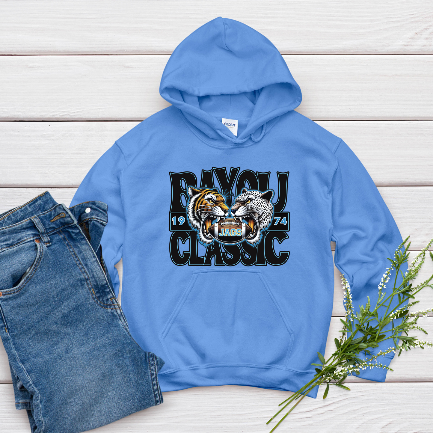 Blue hoodie with a graphic design on a wooden surface with jeans and flowers.  Southern Jags Bayou Classic Apparel - Tailgating T-Shirt, Hoodie & Sweater