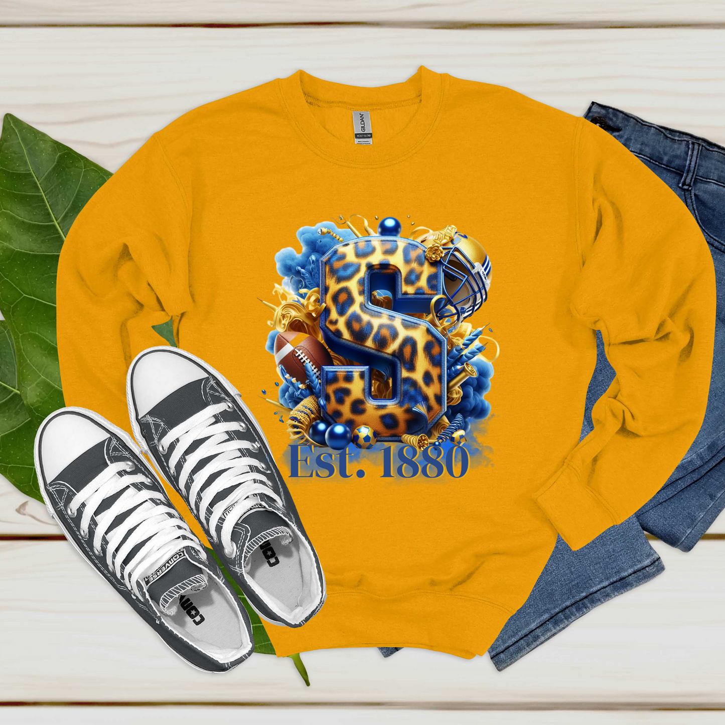 A gold crewneck sweatshirt laid flat on a light wood surface, featuring a large graphic with a leopard-print letter S, football helmet, football, blue paint splatters, and the text 'Est. 1880'.