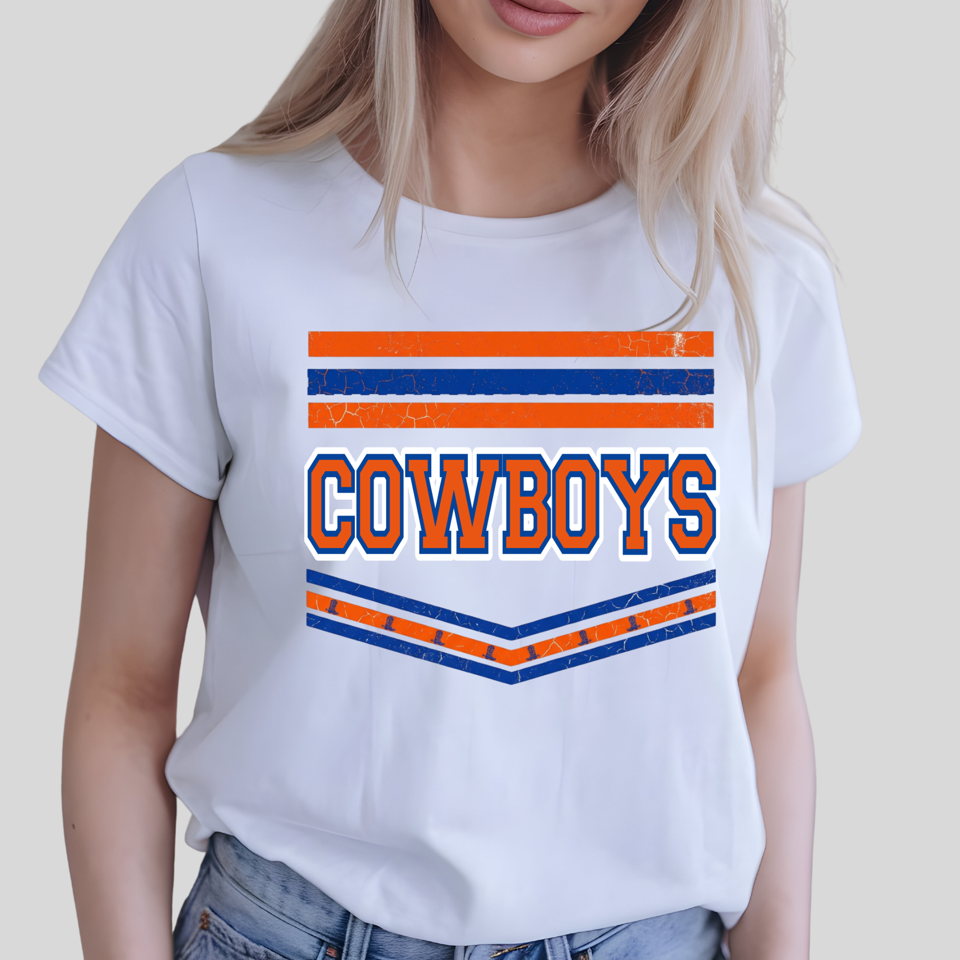 A woman wearing a white short-sleeve t-shirt with a distressed orange and blue 'COWBOYS' graphic on the front, paired with blue jeans, set against a plain light gray background.