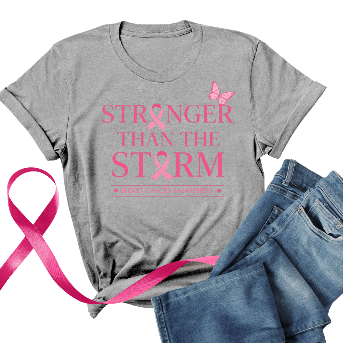 A heather gray short-sleeve T-shirt laid flat with a colorful graphic of a woman in a pink hat and the words 'CRUSH Breast Cancer.' A pink ribbon and blue jeans are placed beside the shirt on a white background.