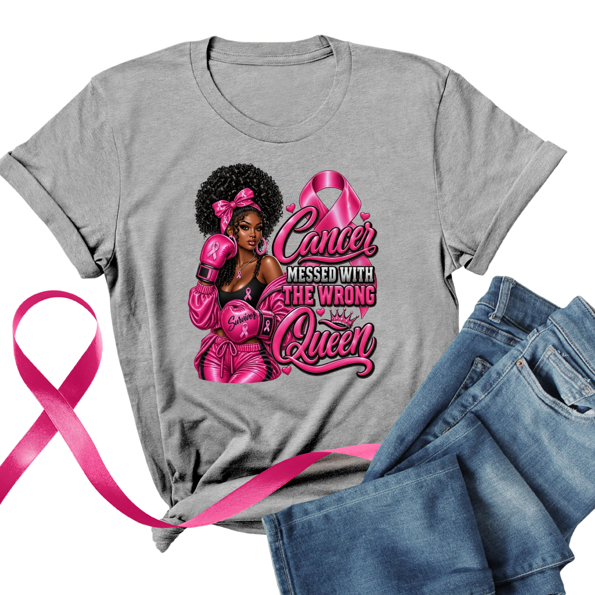 A heather gray short-sleeve graphic T-shirt laid flat with blue jeans beside it and a bright pink ribbon across the lower left. The shirt features a large front print of a Black woman in pink boxing gear with the text 'Cancer Messed With The Wrong Queen' and pink boxing gloves and a pink ribbon.