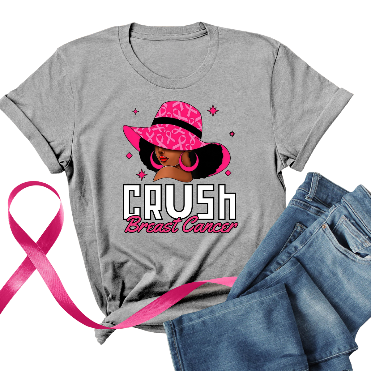 A heather gray short-sleeve T-shirt laid flat with a colorful graphic of a woman in a pink hat and the words 'CRUSH Breast Cancer.' A pink ribbon and blue jeans are placed around it on a white background.