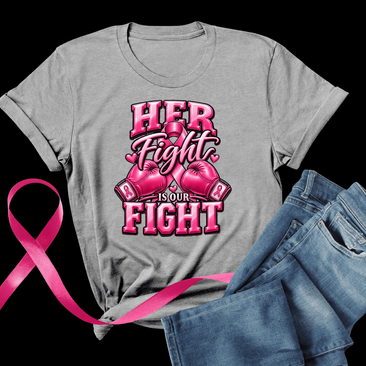 A heather gray short-sleeve T-shirt laid flat on a black background, featuring a pink graphic that reads “HER Fight IS OUR FIGHT” with pink boxing gloves and a pink awareness ribbon. Blue jeans and a pink ribbon are styled around the shirt.