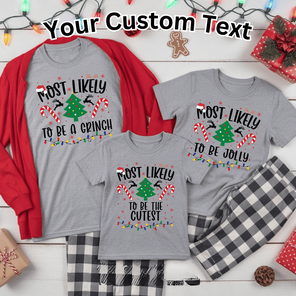 Three gray family Christmas shirts in different sizes displayed on a white wood background with holiday decorations. The shirts feature a custom text graphic that reads 'Most Likely To Be a Grinch / Most Likely To Be Jolly / Most Likely To Be the Cutest'.