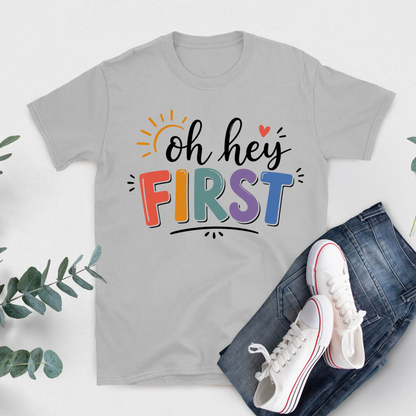 A light gray short-sleeve T-shirt with a colorful 'Kinder Garten' graphic and pink ribbon design, styled in a flat lay with blue jeans, white sneakers, and green eucalyptus leaves on a white background.