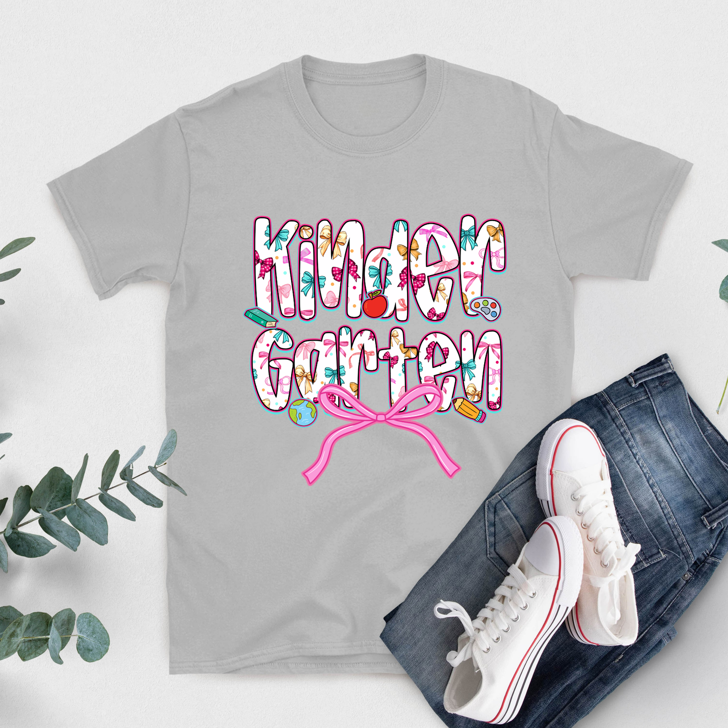 A light gray short-sleeve t-shirt with a colorful 'Kinder Garten' graphic and a pink ribbon, styled in a flat lay with blue jeans, white sneakers, and green eucalyptus leaves on a white background.