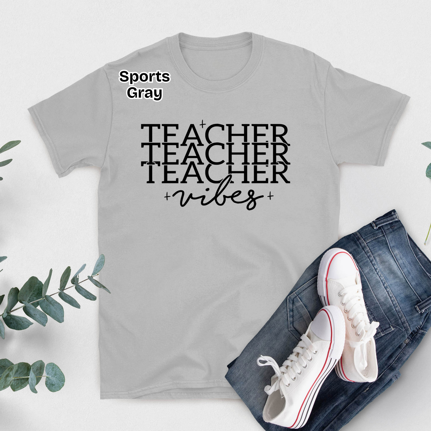 A light gray graphic T-shirt laid flat with black text reading 'TEACHER TEACHER TEACHER vibes' and a small 'Sports Gray' label near the collar. The shirt is styled with blue jeans and white sneakers on a white background with decorative green leaves.