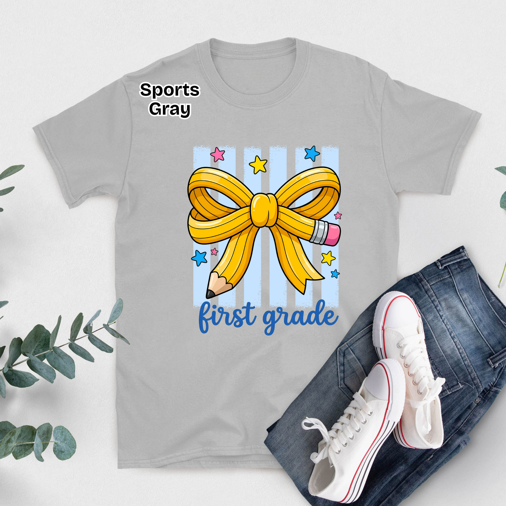 A light gray graphic T-shirt laid flat with blue jeans and white sneakers beside it on a white surface. The shirt has a large yellow ribbon and pencil graphic with blue stripes, stars, and the words 'Sports Gray' and 'first grade'.