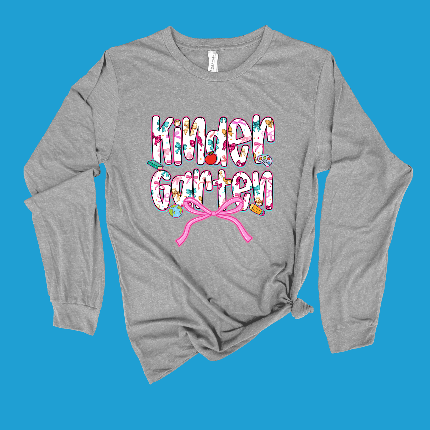 A heather gray long-sleeve t-shirt with a 'Kinder Garten' graphic on the front, featuring colorful letters and a pink ribbon motif, displayed flat against a solid blue background.