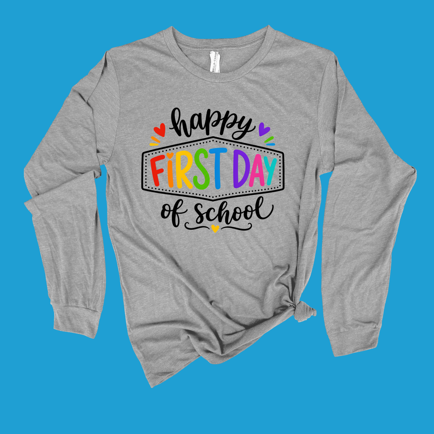 A heather gray long-sleeve t-shirt with a colorful 'happy FIRST DAY of school' graphic on the front, displayed flat against a solid blue background.