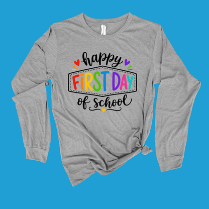 A heather gray long-sleeve t-shirt with a colorful 'happy FIRST DAY of school' graphic on the front, displayed flat against a solid blue background.