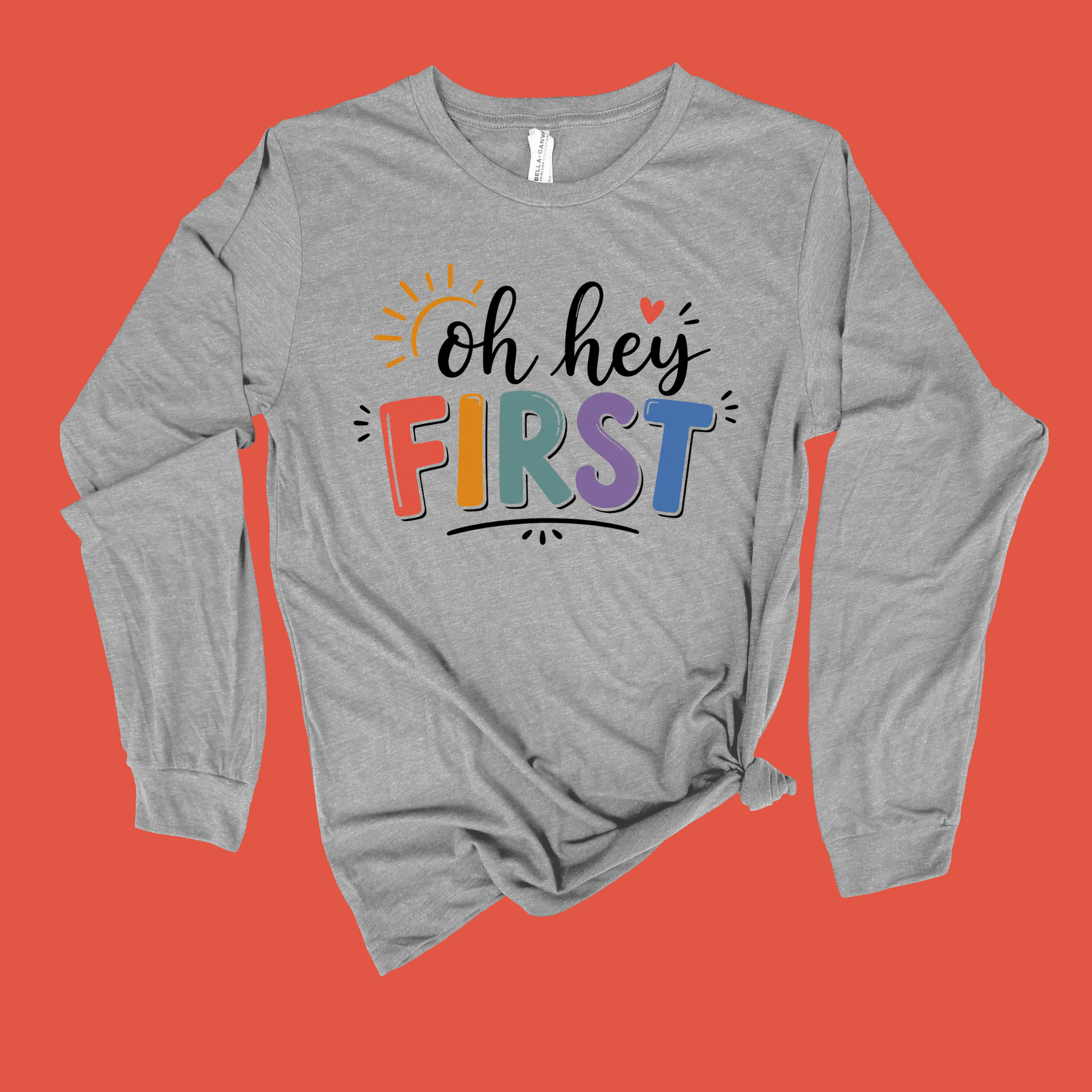A heather gray long-sleeve t-shirt with a 'Kinder Garten' graphic on the front, featuring a pink ribbon and small colorful illustrations, displayed flat against a solid blue background.