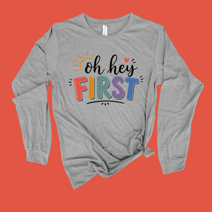 A heather gray long-sleeve t-shirt with a 'Kinder Garten' graphic on the front, featuring a pink ribbon and small colorful illustrations, displayed flat against a solid blue background.