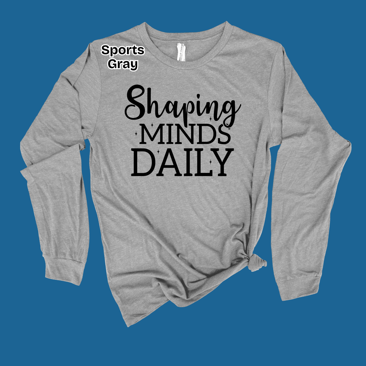 A heather gray long-sleeve t-shirt with black text reading 'Shaping MINDS DAILY' on the front, styled with a knot at the bottom hem and a small 'Sports Gray' label near the collar, set against a solid blue background.