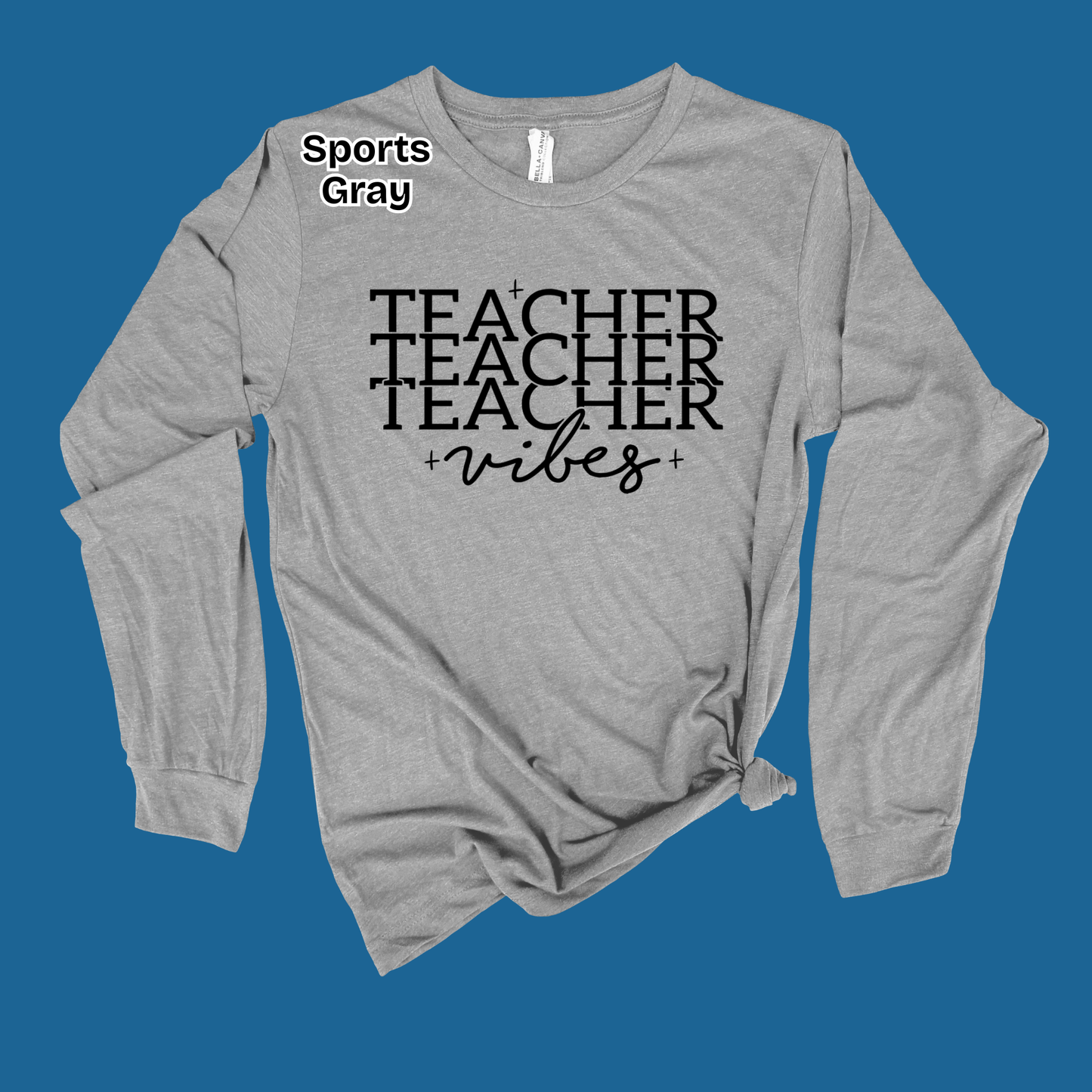 A heather gray long-sleeve t-shirt with a black graphic text design reading 'TEACHER TEACHER TEACHER vibes' on the front, styled with a knot at the bottom hem and a small 'Sports Gray' label near the collar, set against a solid blue background.