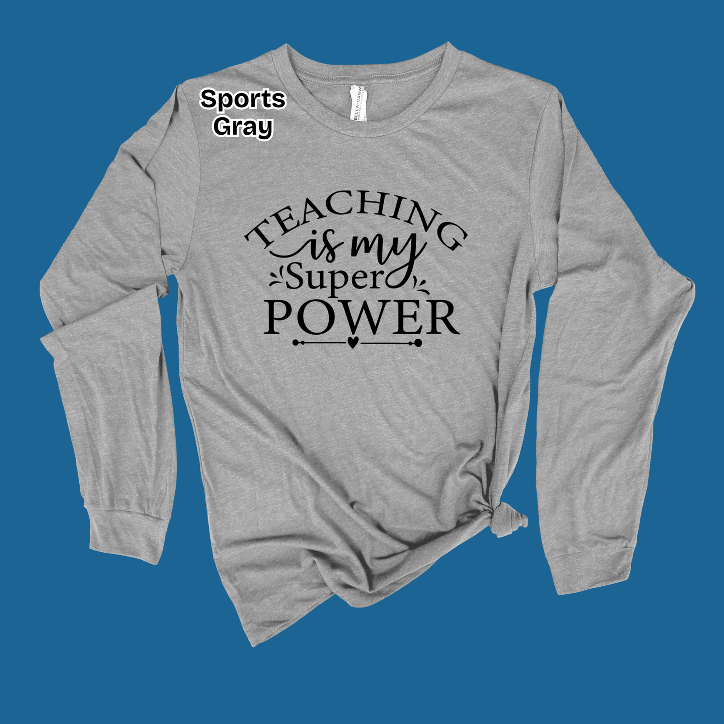 A heather gray long-sleeve t-shirt with a black graphic text print reading 'TEACHING is my Super POWER' on the front, styled with a knot at the bottom hem against a solid blue background.