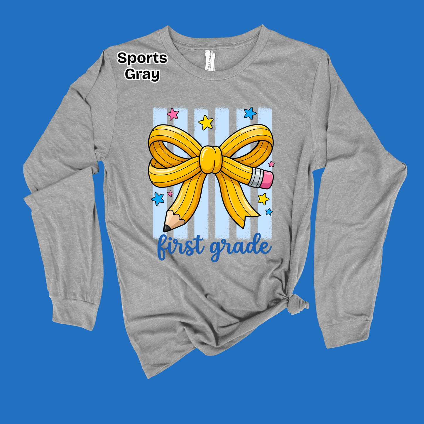 A heather gray long-sleeve t-shirt with a 'first grade' first birthday graphic, featuring a yellow ribbon bow, pencil, and stars on a blue striped background, displayed on a solid blue backdrop.