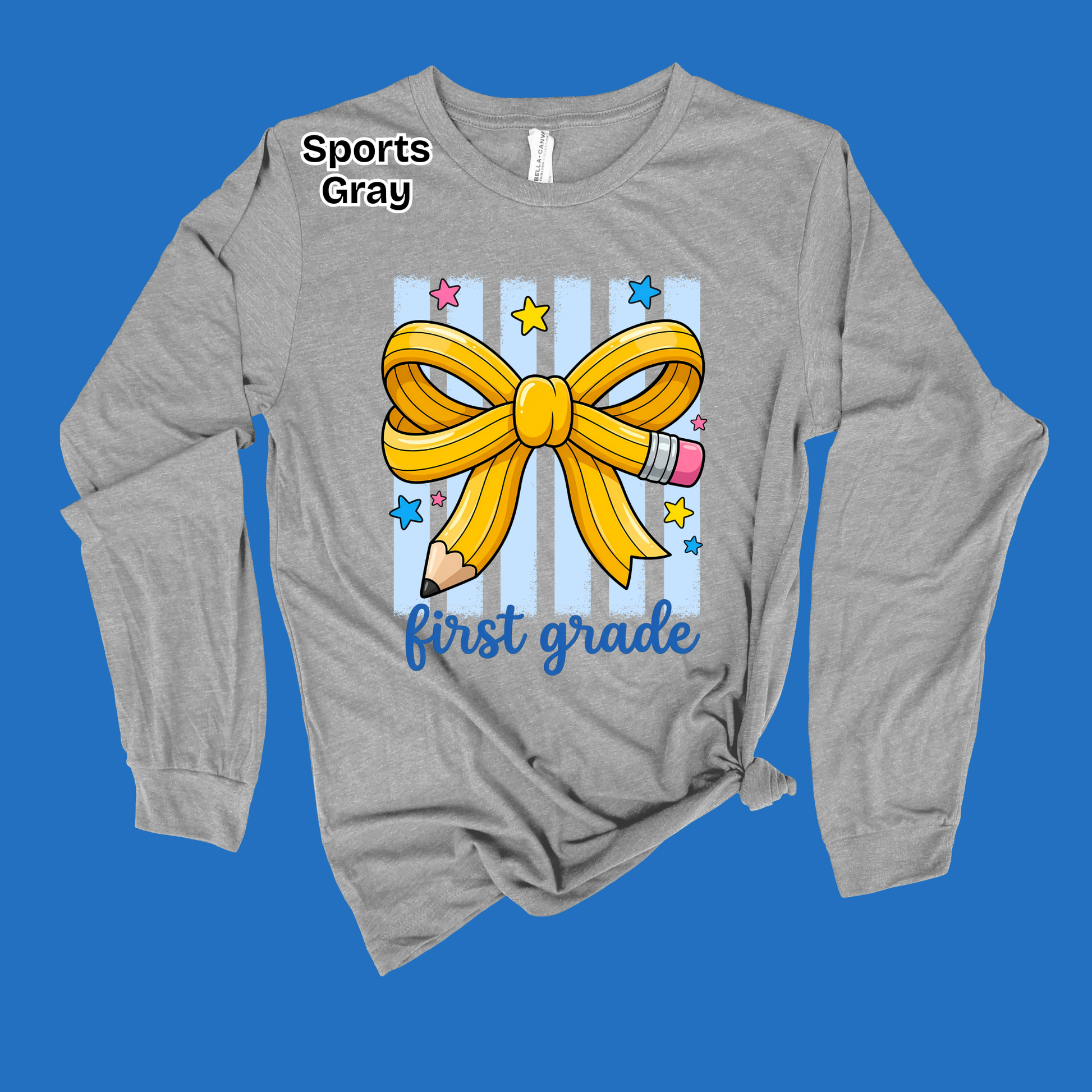 A heather gray long-sleeve t-shirt with a 'first grade' first birthday graphic, featuring a yellow ribbon bow, pencil, and stars on a blue striped background, displayed on a solid blue backdrop.