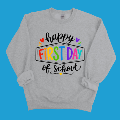 A light heather gray crewneck sweatshirt with a colorful graphic that reads 'happy FIRST DAY of school' in black script and rainbow-colored block letters, displayed on a solid blue background.