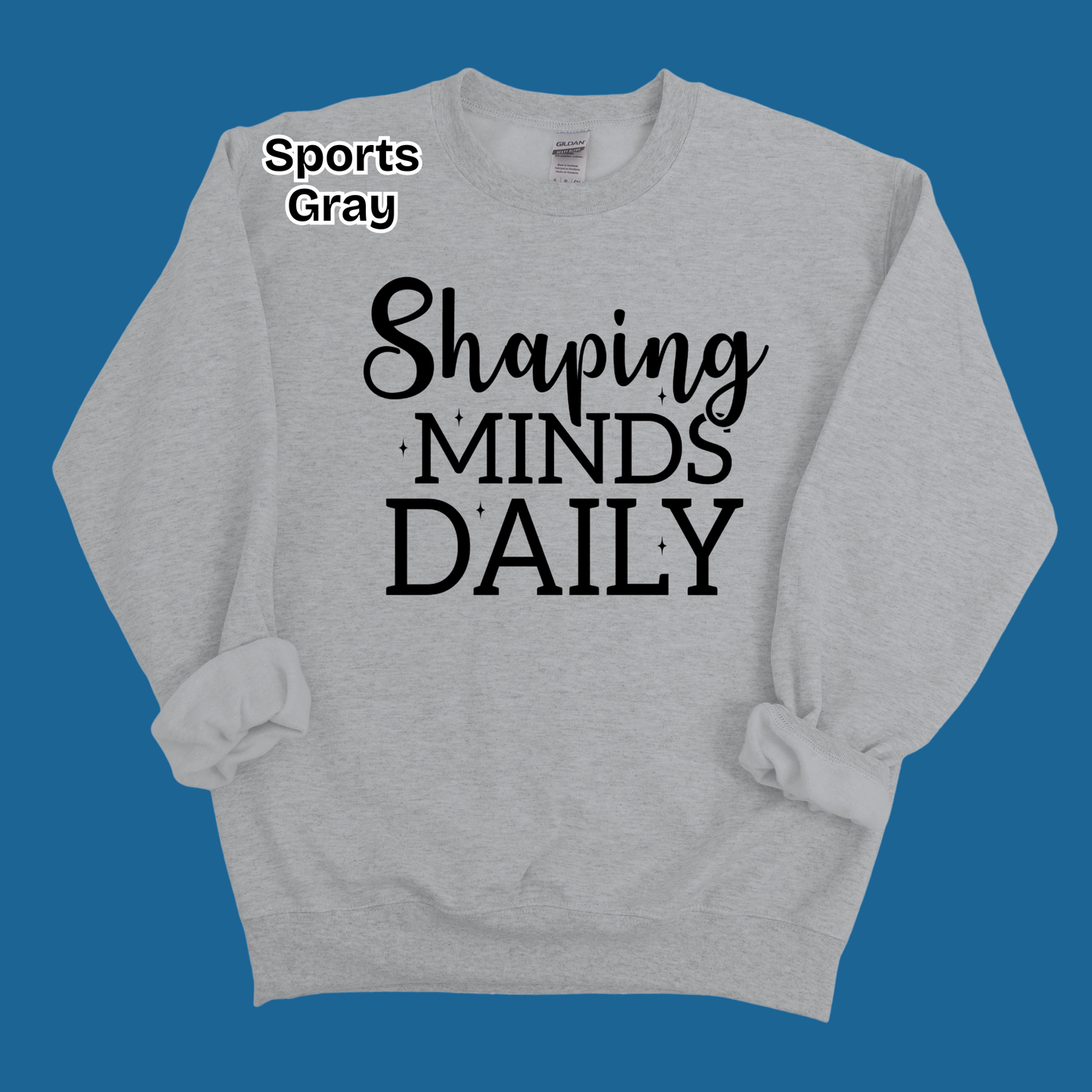 A light gray crewneck sweatshirt with rolled cuffs, featuring the text 'Shaping MINDS DAILY' in black script and block lettering on the front. The text 'Sports Gray' is printed in white on the upper left chest.