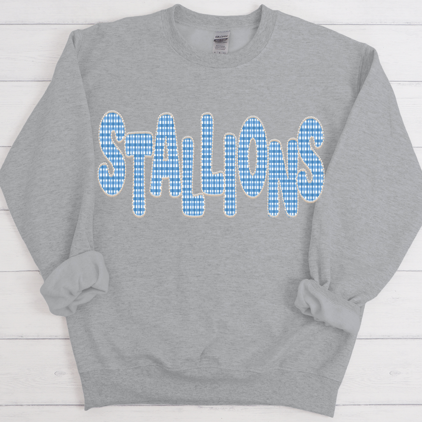A heather gray crewneck sweatshirt featuring the word 'STALLIONS' printed across the chest in a blue and white gingham pattern with a white outline, laid flat on a white wood plank background.
