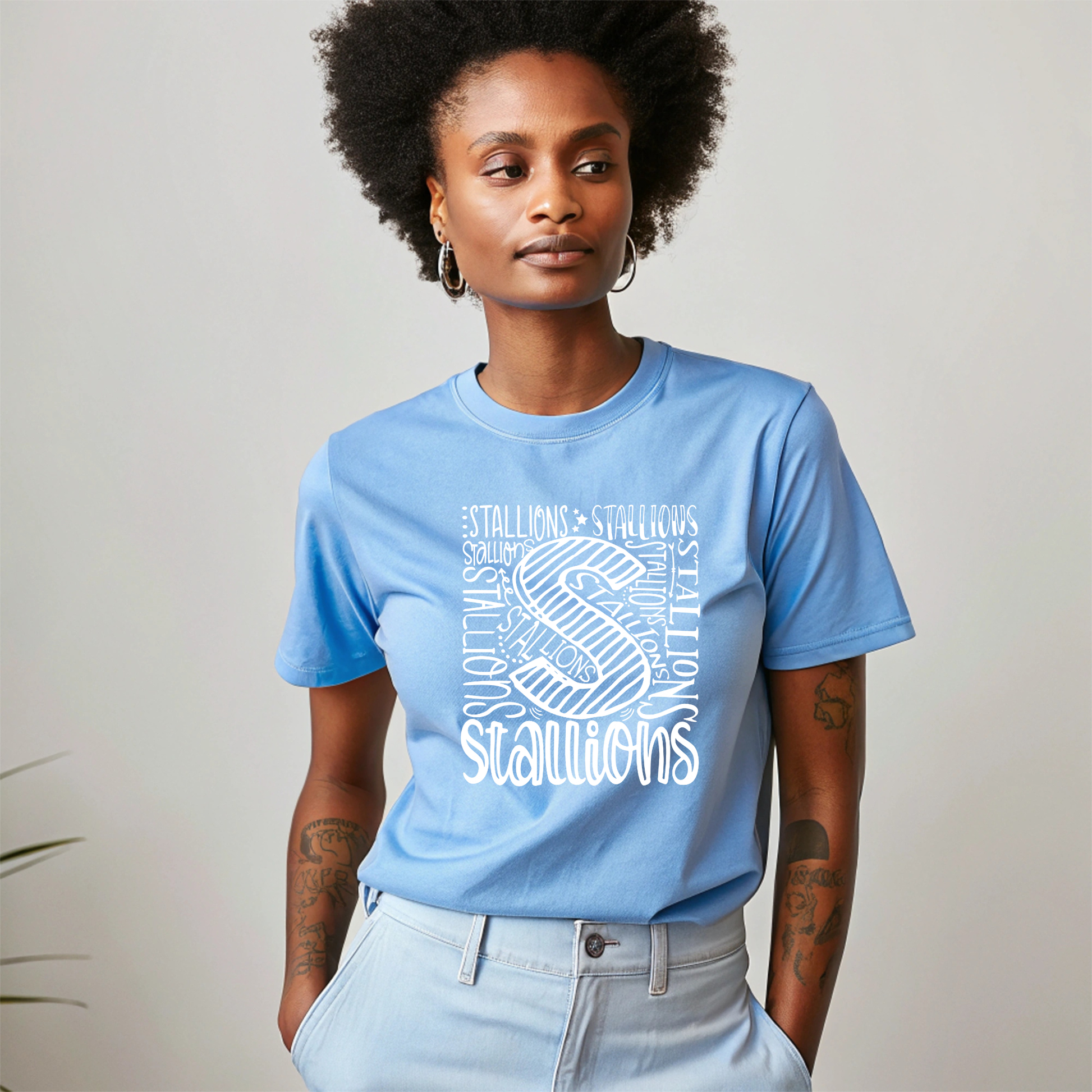 A woman wearing a light blue crewneck t-shirt with a white graphic print of a horse and the word 'Stallions' repeated in the design, paired with light-wash jeans against a neutral studio background.