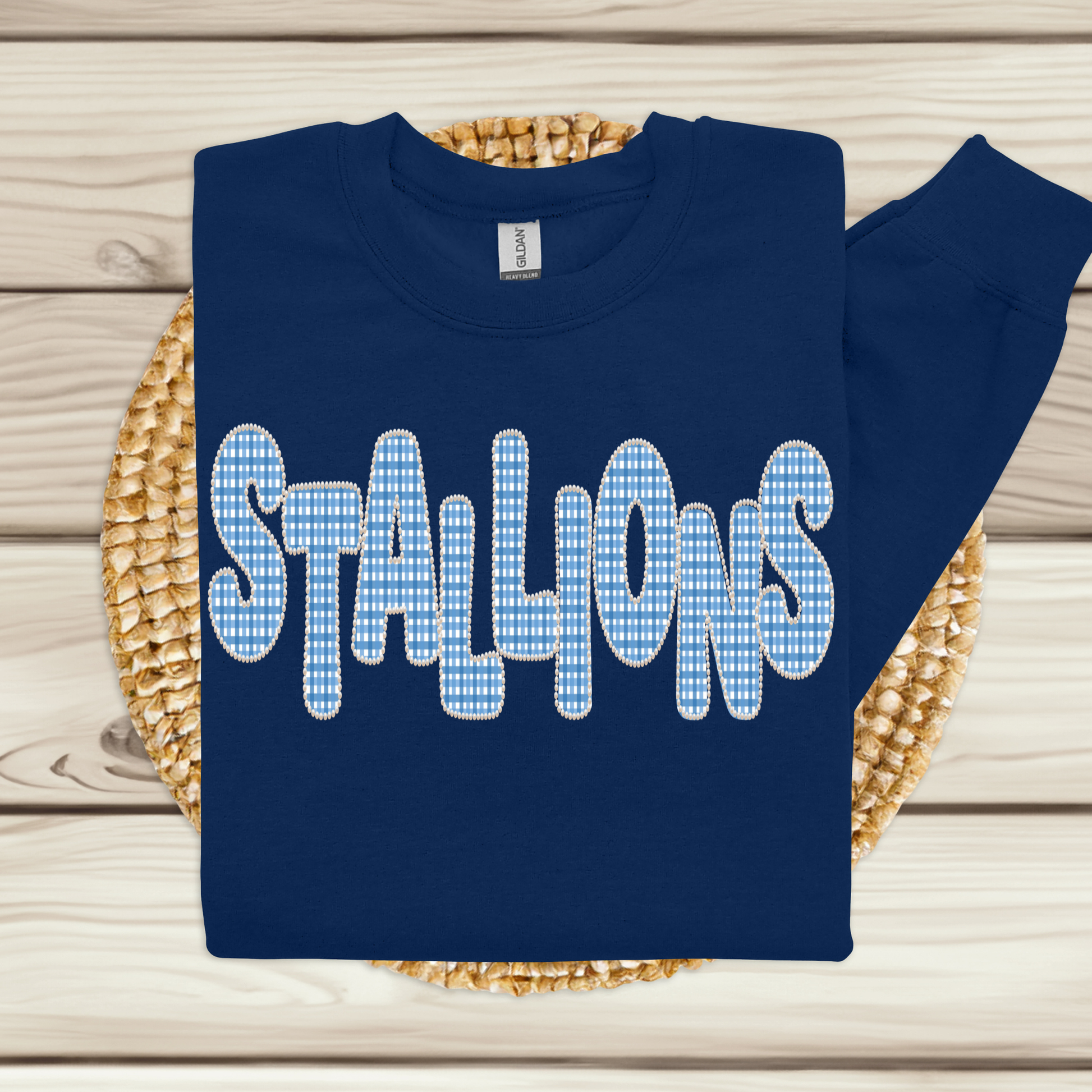 A navy blue sweatshirt with the word 'STALLIONS' printed across the chest in large white letters filled with a light blue and white gingham check pattern, displayed on a woven straw mat over a light wood plank background.