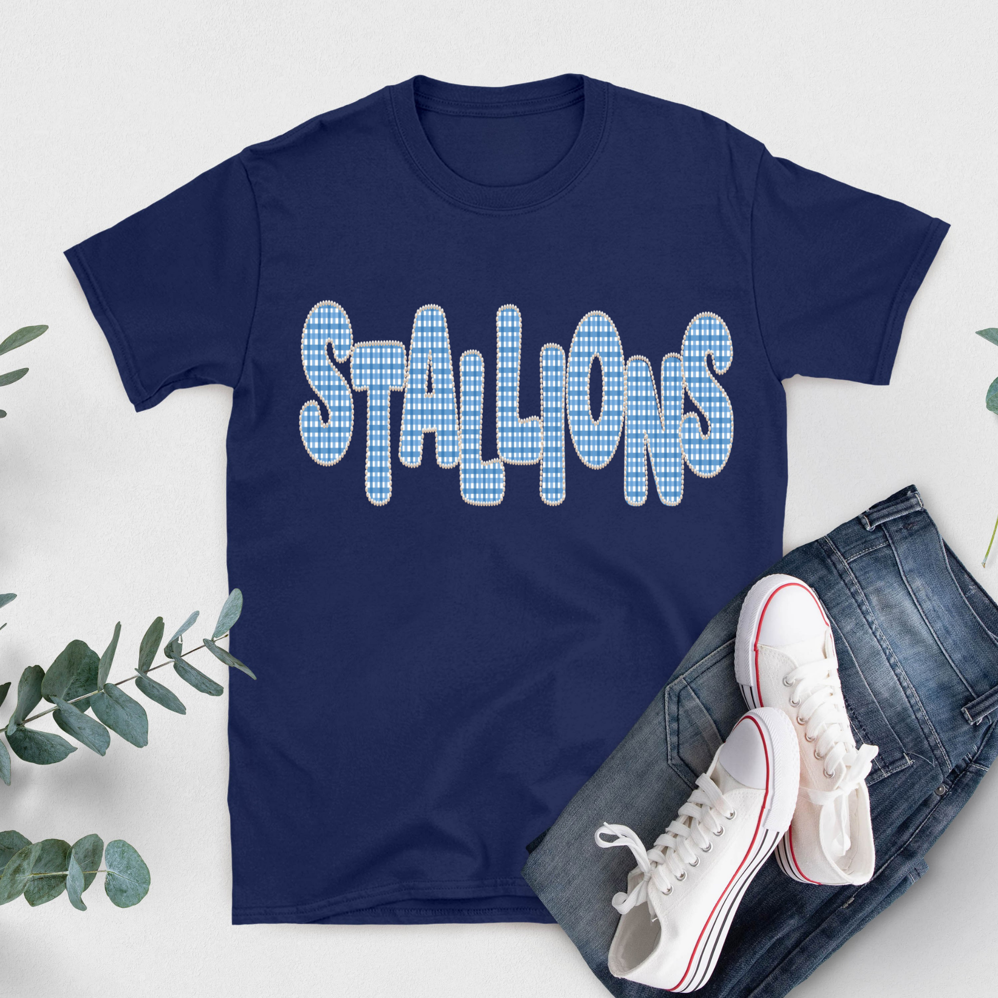 A navy blue short-sleeve t-shirt with the word 'STALLIONS' printed across the chest in a light blue and white gingham pattern. The shirt is styled in a flat lay with blue jeans, white sneakers, and green eucalyptus leaves on a light gray background.