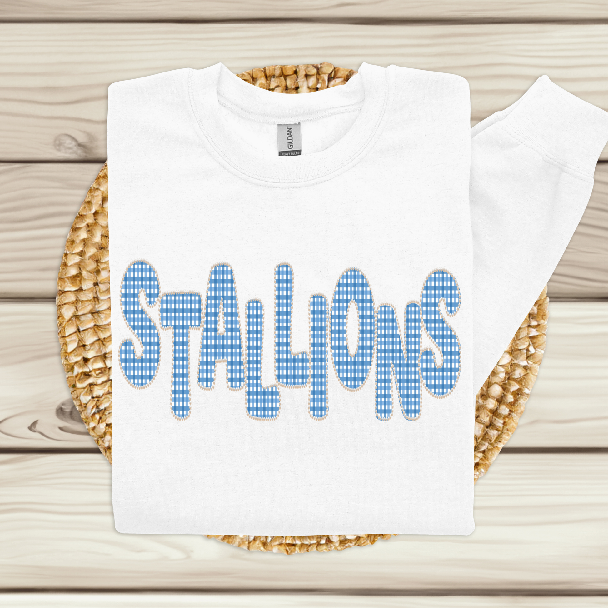 A white crewneck sweatshirt with the word 'STALLIONS' printed across the chest in a blue-and-white gingham patterned font, displayed on a woven round mat over a light wood plank background.