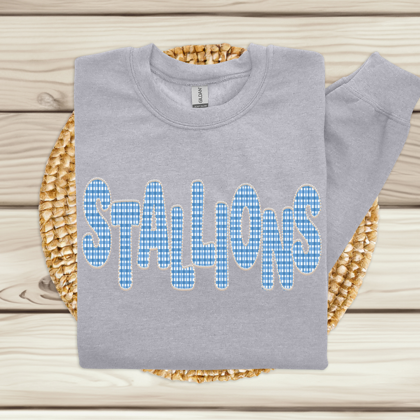 A light gray sweatshirt laid flat on a woven round mat over a light wood plank surface. The sweatshirt has large blue-and-white gingham patterned letters spelling "STALLIONS" across the front.