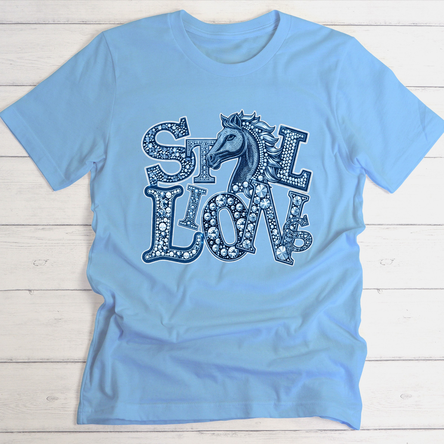A light blue short-sleeve t-shirt with a decorative graphic on the front featuring the words 'SILK LION' in a patterned font with a horse head integrated into the design, laid flat on a white wood plank background.