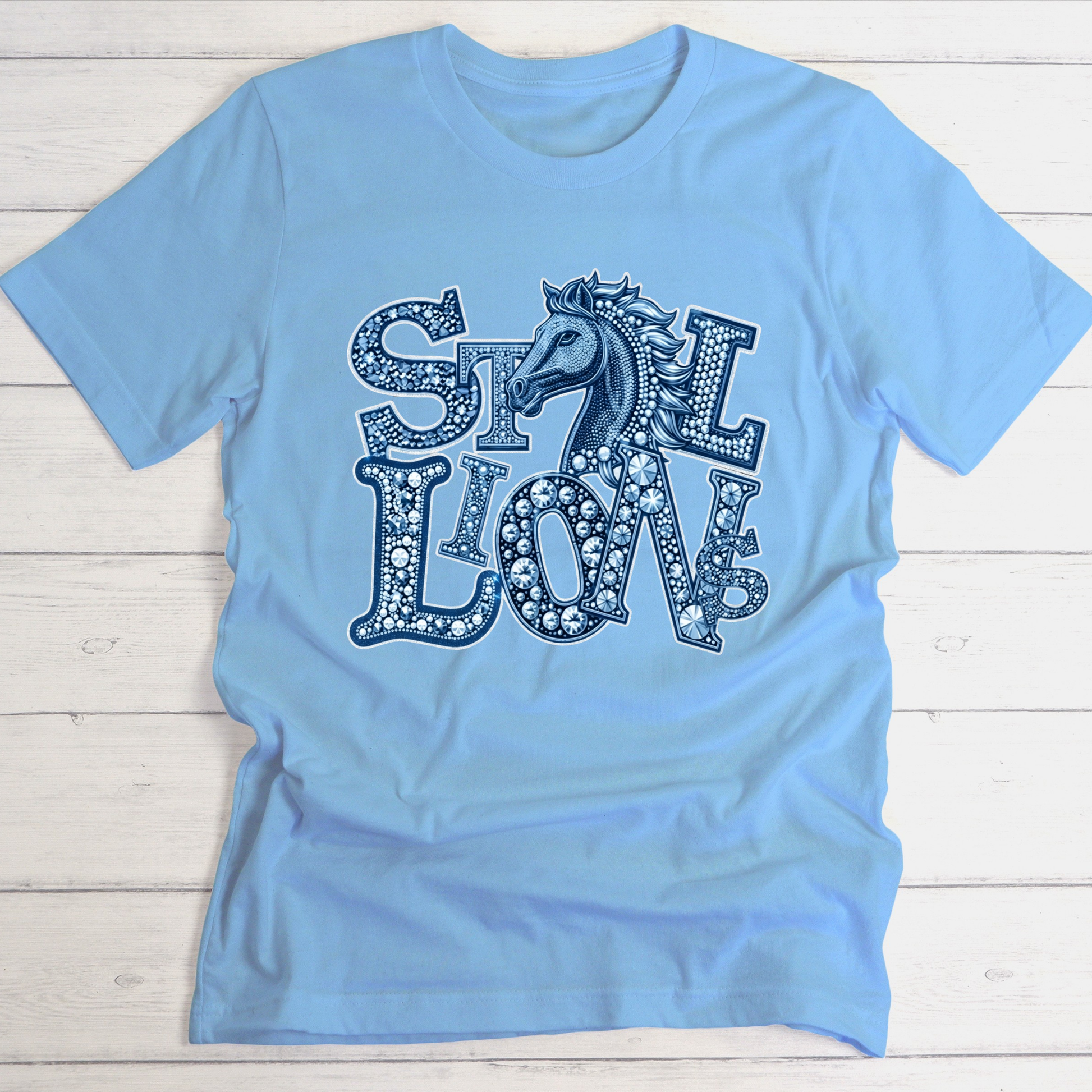 A light blue short-sleeve t-shirt with a decorative graphic on the front featuring the words 'SILK LION' in a patterned font with a horse head integrated into the design, laid flat on a white wood plank background.
